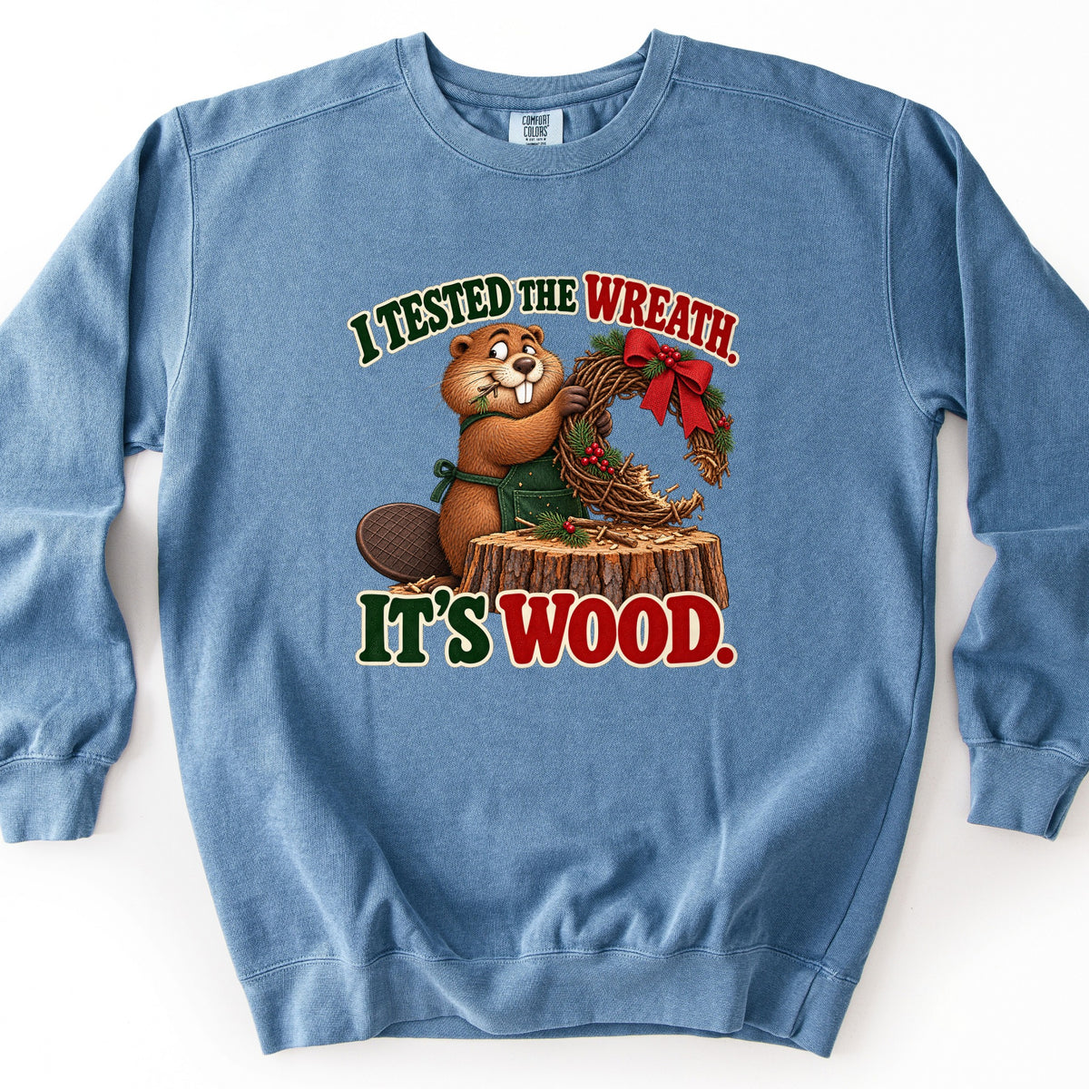 I Tested the Wreath. It's Wood. Comfort Colors crewneck sweatshirt in Blue Jean, laid flat. A cartoon beaver in a green apron holds a half-chewed twig wreath with a red bow and berries on a tree stump, between the words I Tested the Wreath. and It's Wood. Laid on a white background.