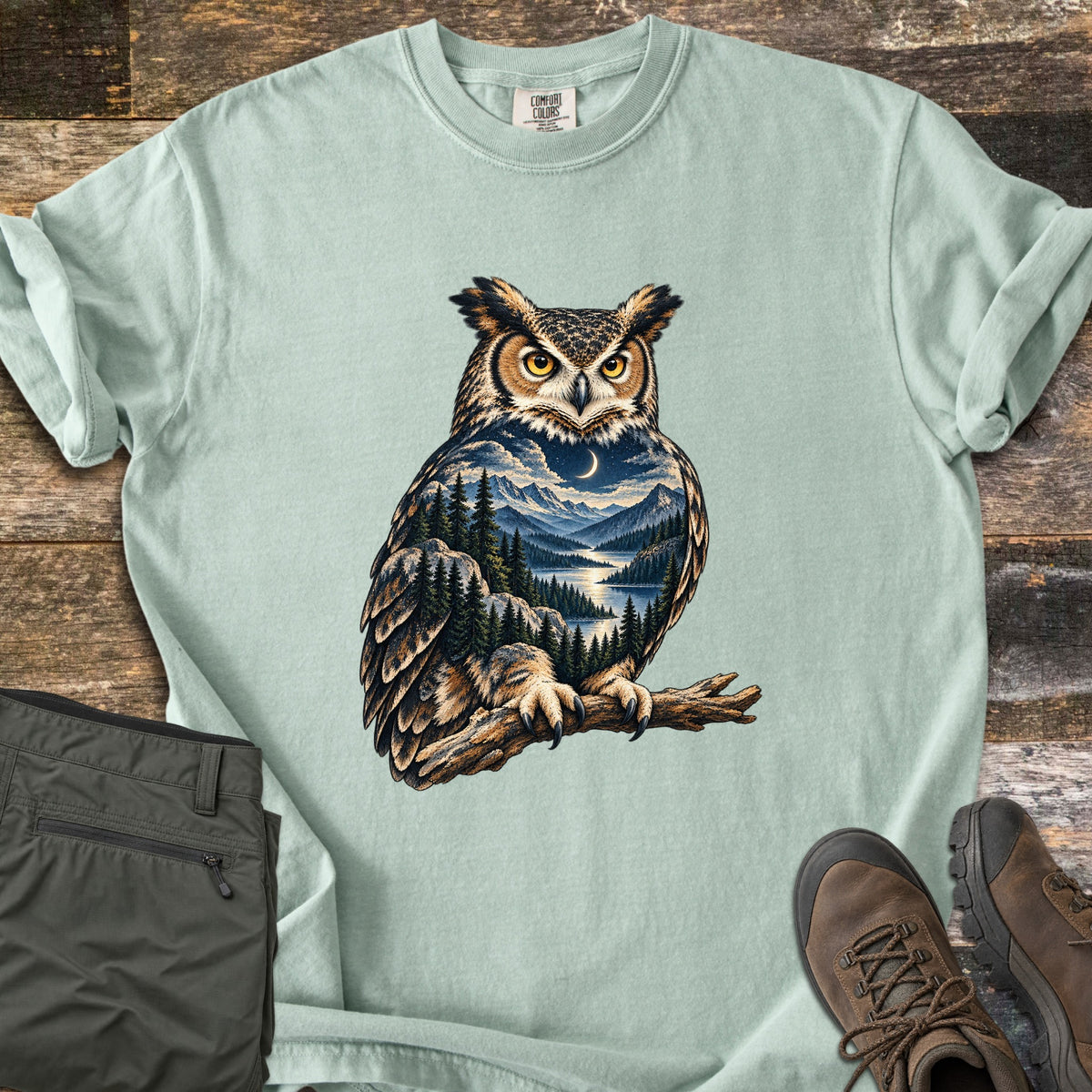 Owl Mountain Lake Comfort Colors T-shirt in Bay, laid flat. A great horned owl perched on a branch, its body filled with a night scene of a crescent moon, snowy mountains, pine forest and a lake. Olive hiking pants and brown boots sit beside it on weathered wood.