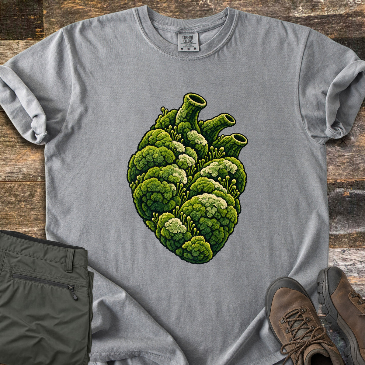 Moss Heart Comfort Colors T-shirt in Grey, laid flat. An anatomical heart covered in green moss with pale tufts, its vessels shaped like moss-covered tubes. The shirt has rolled sleeves and lies on weathered wood beside folded olive pants and brown hiking boots.