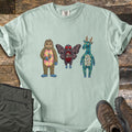 Patchwork Cryptid Trio Comfort Colors T-shirt in Bay, laid flat. Three patchwork fabric cryptids: Bigfoot with a pink argyle belly holding a footprint patch, a red-eyed Mothman, and a plaid jackalope. Laid on weathered wood beside olive pants and brown hiking boots.