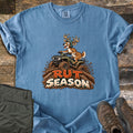 Rut Season Comfort Colors T-shirt in Blue Jean, laid flat. A cartoon buck in a green helmet rides a four-wheeler through splashing mud above the words Rut Season. Sleeves are rolled on a wood plank surface beside folded olive pants and brown hiking boots.
