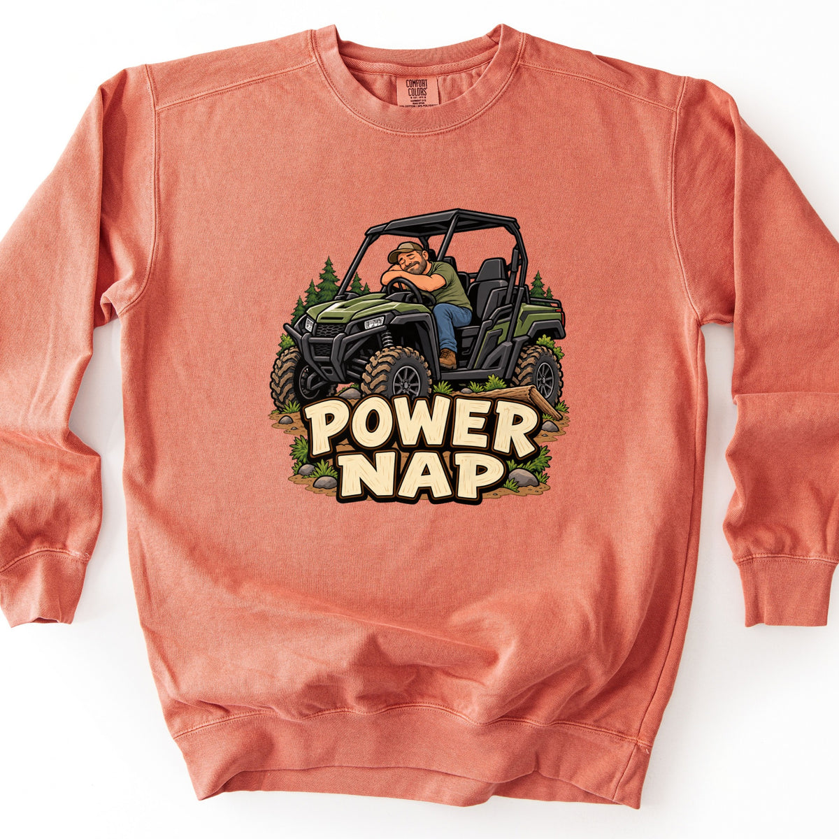 Power Nap Comfort Colors crewneck sweatshirt in Terracotta, laid flat. A cartoon bearded man in a cap naps slumped over the steering wheel of a green side-by-side UTV among pine trees and mud, above the words Power Nap in cream letters. Shown on a white background.