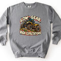 Low Gear High Expectations Comfort Colors crewneck sweatshirt in Grey, laid flat. A rider in a teal helmet and jacket drives a mud-splattered yellow ATV over a dirt mound with grass, between the words Low Gear and High Expectations in cream lettering. Shown on a white background.