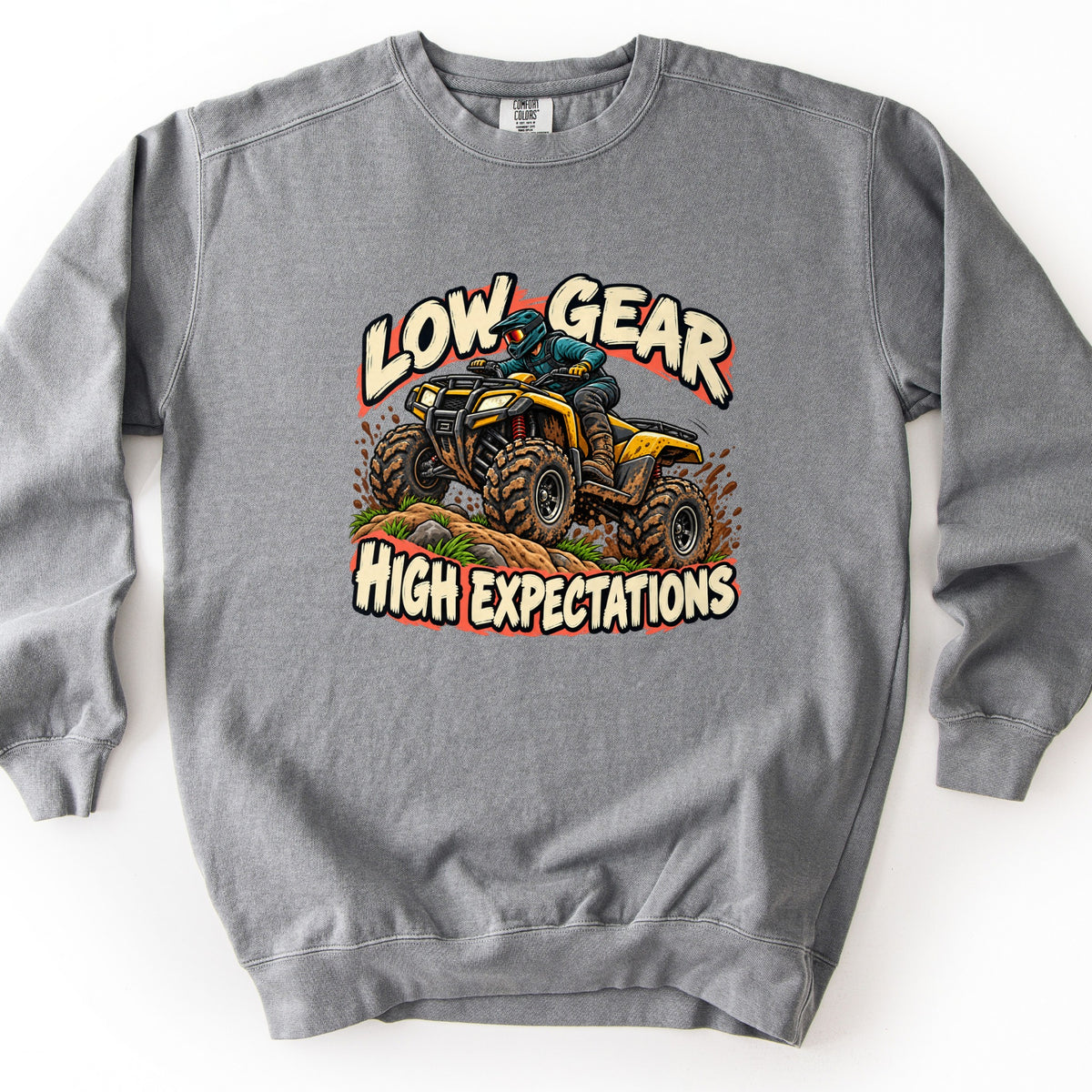 Low Gear High Expectations Comfort Colors crewneck sweatshirt in Grey, laid flat. A rider in a teal helmet and jacket drives a mud-splattered yellow ATV over a dirt mound with grass, between the words Low Gear and High Expectations in cream lettering. Shown on a white background.