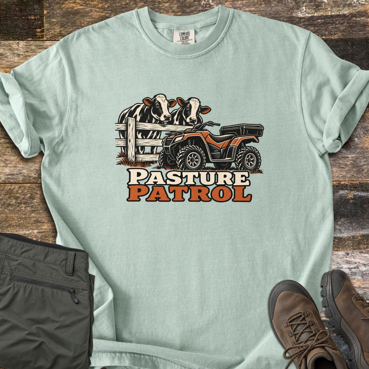 Pasture Patrol Comfort Colors T-shirt in Bay, laid flat. Two black and white cows lean over a wooden fence beside an orange and black ATV, above the words Pasture Patrol. Folded olive pants and brown hiking boots sit on a weathered wood surface.