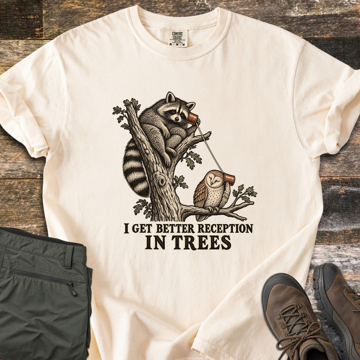 I Get Better Reception in Trees Comfort Colors T-shirt in Ivory, laid flat. A vintage-style sketch of a raccoon in a tree holding a tin can phone, connected by string to a barn owl on a branch below, above the words I Get Better Reception In Trees. Olive pants and brown hiking boots sit on weathered wood.
