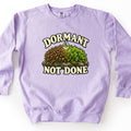 Dormant Not Done Comfort Colors crewneck sweatshirt in Orchid, laid flat. A water droplet falls onto a clump of moss on a rock, dry and brown on the left and bright green on the right, between the words DORMANT and NOT DONE in cream lettering. It lies on a white surface.