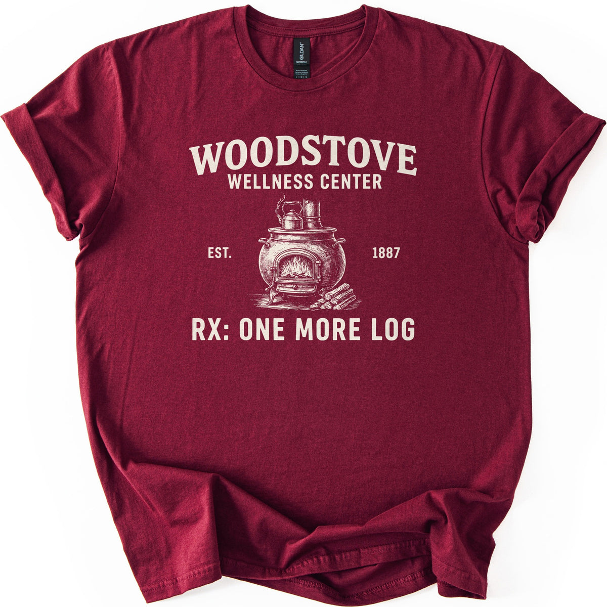 Woodstove Wellness Center Comfort Colors T-shirt in Maroon, laid flat. A cream sketch of a round woodstove with a kettle on top, flames in its open door and stacked logs beside it, under Woodstove Wellness Center, Est. 1887, and Rx: One More Log. Sleeves rolled, on white.