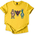 Patchwork Cryptid Trio Comfort Colors T-shirt in Daisy, laid flat. Three patchwork fabric cryptids with no words: Bigfoot with a pink argyle belly holding a footprint, Mothman with red button eyes, and an antlered teal plaid creature. Sleeves cuffed, on white.