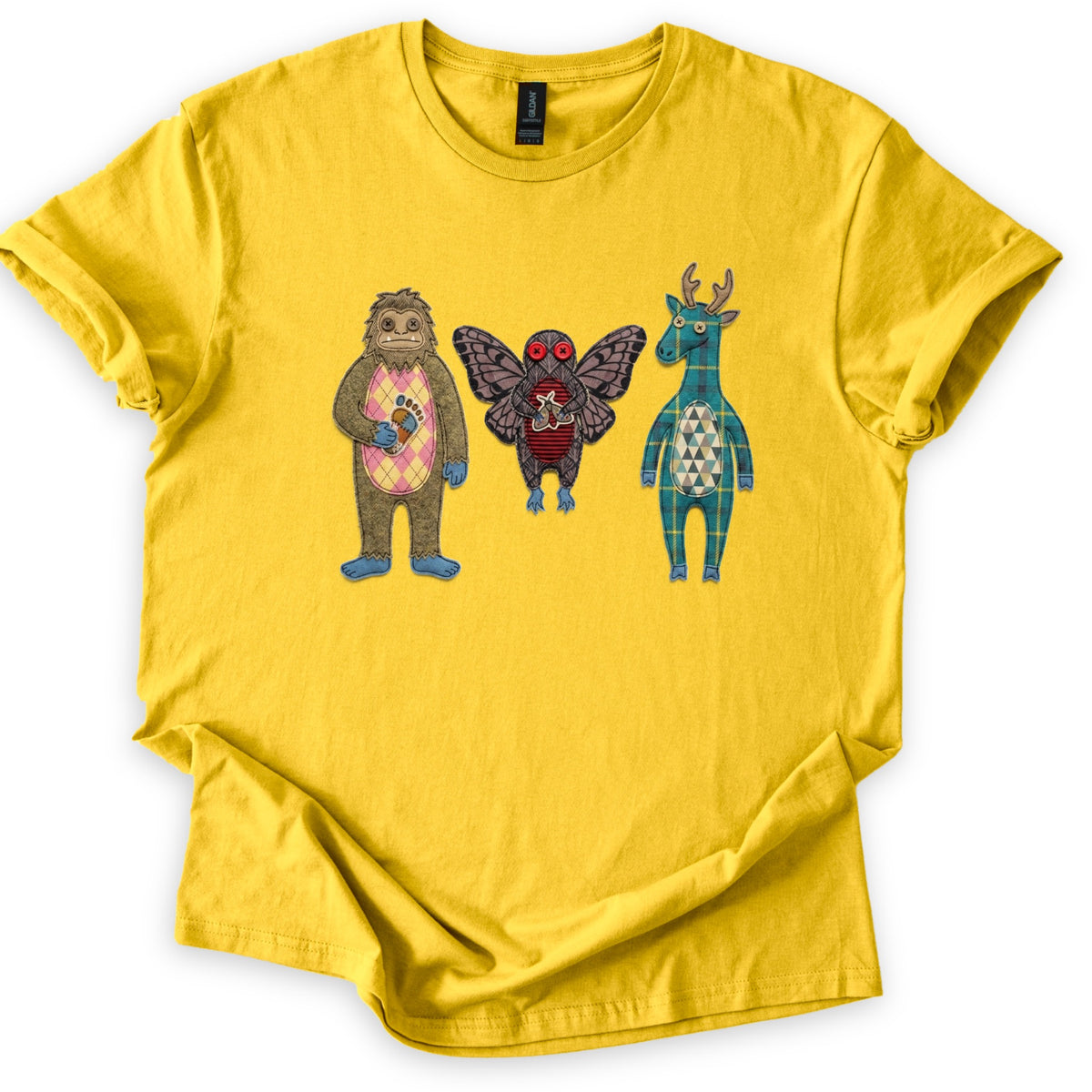Patchwork Cryptid Trio Comfort Colors T-shirt in Daisy, laid flat. Three patchwork fabric cryptids with no words: Bigfoot with a pink argyle belly holding a footprint, Mothman with red button eyes, and an antlered teal plaid creature. Sleeves cuffed, on white.