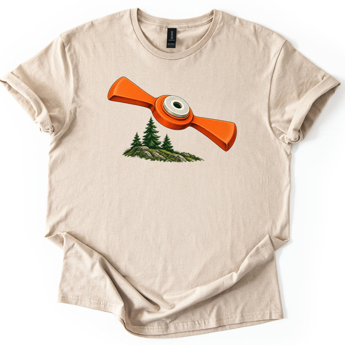 Winged Target Over the Pines Comfort Colors T-shirt in Sand, laid flat. An orange winged clay target with a white center ring flies above three pine trees on a mossy, rocky mound, with no words. The shirt lies on a white surface with its sleeves cuffed.
