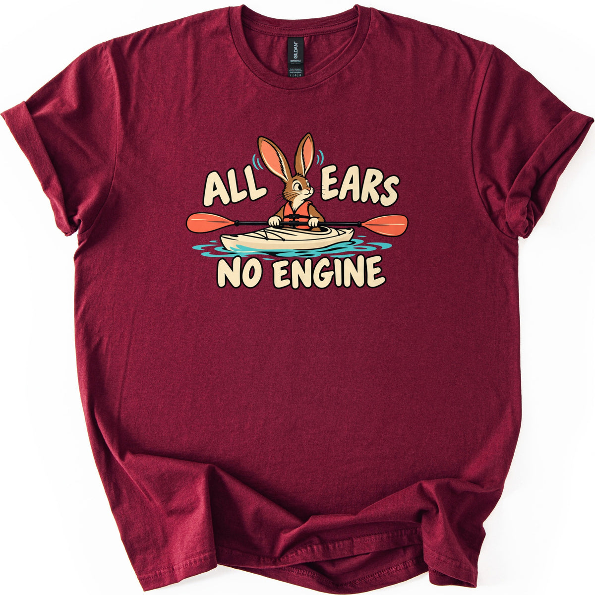 All Ears No Engine Comfort Colors T-shirt in Maroon, laid flat. A cartoon rabbit in an orange life vest paddles a cream kayak on blue water, its tall ears between the words All Ears, with No Engine below. The shirt has rolled sleeves on a white background.