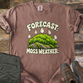 Forecast Moss Weather Comfort Colors T-shirt in Espresso, laid flat. A cartoon mound of bright green moss on a gray rock, with raindrops falling from the words Forecast: above and Moss Weather. below. Olive hiking pants and brown hiking boots sit beside it on weathered wood boards.