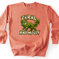 Feral and Mossy Comfort Colors crewneck sweatshirt in Terracotta, laid flat. A cartoon cat made of dripping green moss with bark patches walks and smiles back over its shoulder. Moss-draped cream letters read FERAL above it and AND MOSSY below. Shown on a white background.