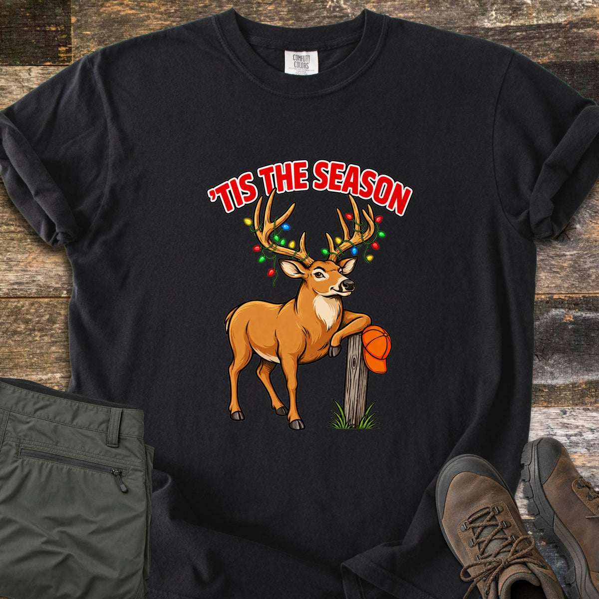 Buck Tis The Season Comfort Colors T-shirt in Black, laid flat. A cartoon buck with colorful Christmas lights wrapped in its antlers leans on a wooden post holding an orange cap, under red words 'Tis The Season. Olive pants and brown hiking boots lie on weathered wood.