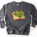 Moss Storybook Comfort Colors crewneck sweatshirt in Pepper, laid flat. An open antique book with a red leather cover has green moss, gray rocks, and tiny sprouting spore capsules growing from its pages, printed in the center front with no words. The sweatshirt lies on a white surface.