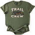 Trail Crew Comfort Colors T-shirt in Military Green, laid flat. Cream distressed letters read TRAIL above and CREW below a black ATV, with an orange bow saw resting on a cut branch and leaves in front of it. The shirt has rolled sleeves on a white background.