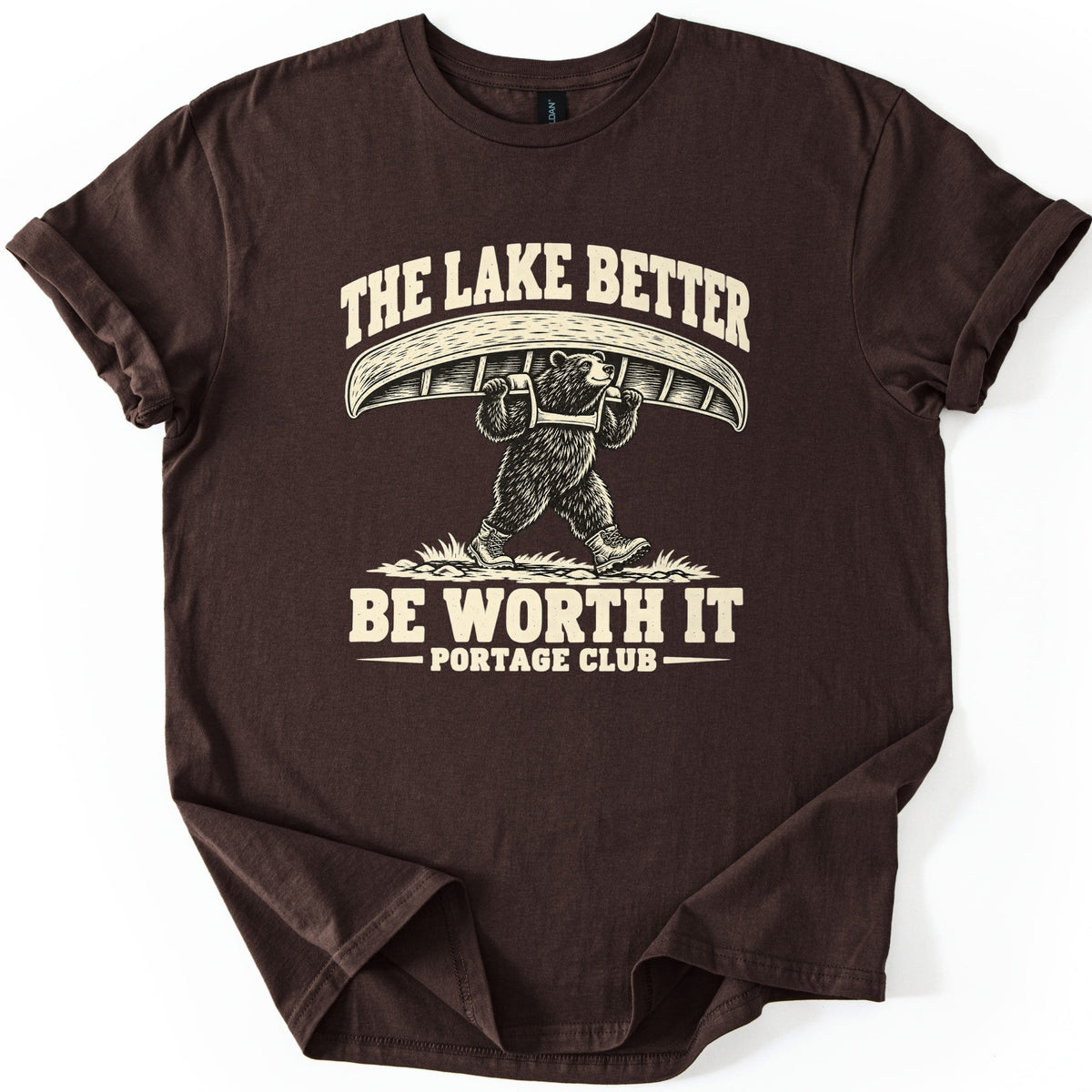 The Lake Better Be Worth It Comfort Colors T-shirt in Dark Chocolate, laid flat. A cream-colored drawing of a bear in hiking boots carrying an overturned canoe on its shoulders, with the words The Lake Better Be Worth It and Portage Club. The sleeves are cuffed on a white background.