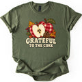 Grateful to the Core Comfort Colors T-shirt in Military Green, laid flat. Two red and cream gingham apples, one sliced in half to show its seeds, sit with a pinecone, orange autumn leaves and red berries above the words Grateful to the Core. Sleeves cuffed, on a white background.