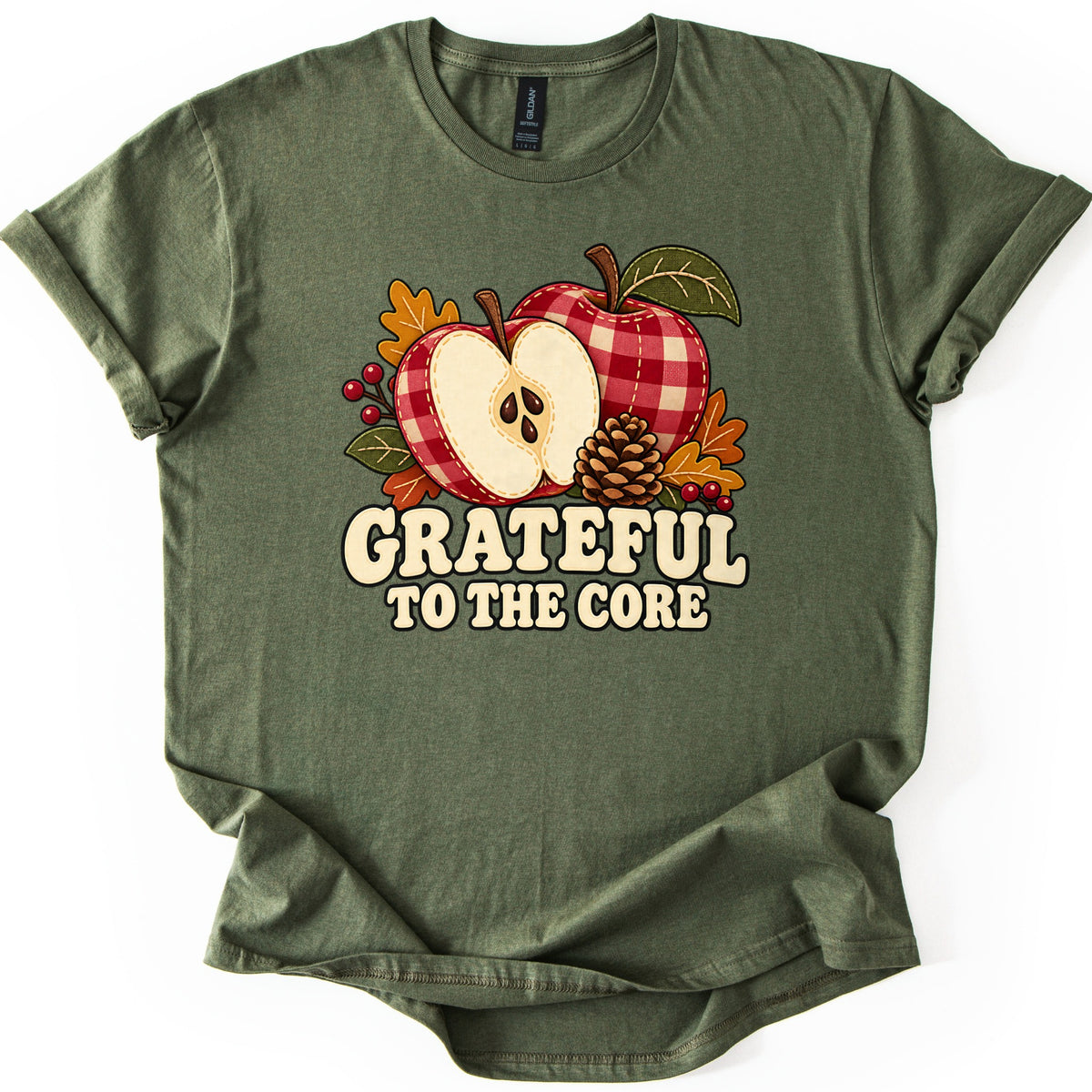 Grateful to the Core Comfort Colors T-shirt in Military Green, laid flat. Two red and cream gingham apples, one sliced in half to show its seeds, sit with a pinecone, orange autumn leaves and red berries above the words Grateful to the Core. Sleeves cuffed, on a white background.