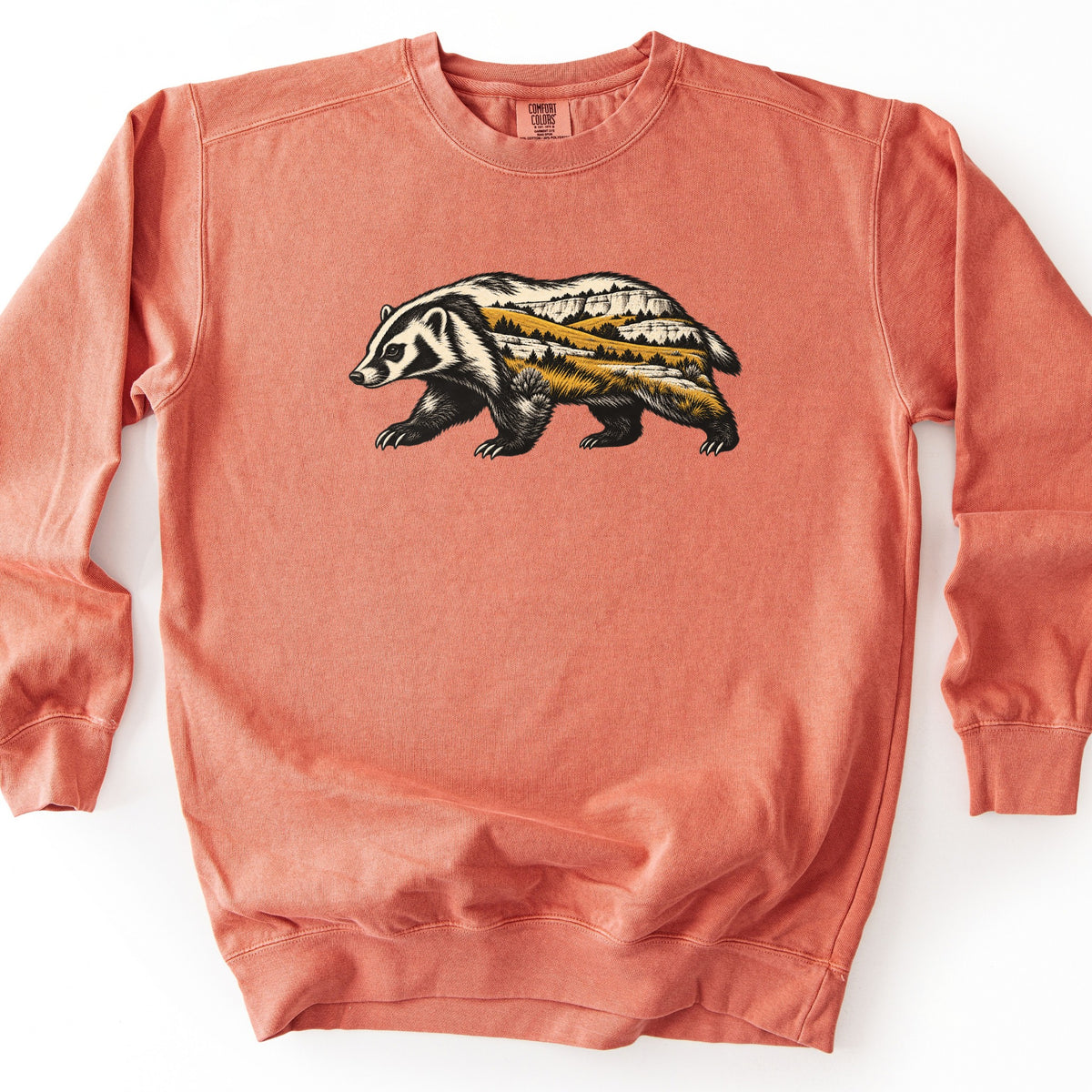 Badger Prairie Bluffs Comfort Colors crewneck sweatshirt in Terracotta, laid flat. A walking badger in black and cream fur, its body filled with a prairie scene of white bluffs, golden grassy hills and dark pine trees. No words are printed. It lies on a plain white surface.