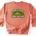 Hide & Seek Moss Edition Comfort Colors crewneck sweatshirt in Terracotta, laid flat. A green, dark-spotted toad peeks out of bright green moss between cream arched letters reading Hide & Seek above and Moss Edition below. The sweatshirt lies on a white background.