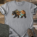 Bear River Canyon Comfort Colors T-shirt in Grey, laid flat. A walking black bear whose body is filled with a canyon scene of orange cliffs, pine trees, a turquoise river and snowy peaks. Folded olive hiking pants and brown hiking boots sit on weathered wood boards.