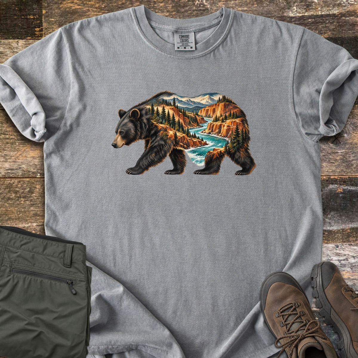 Bear River Canyon Comfort Colors T-shirt in Grey, laid flat. A walking black bear whose body is filled with a canyon scene of orange cliffs, pine trees, a turquoise river and snowy peaks. Folded olive hiking pants and brown hiking boots sit on weathered wood boards.