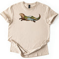 Otter River Bend Comfort Colors T-shirt in Sand, laid flat. A river otter stretches forward with its head raised, its body filled with a stream rushing over boulders, pine trees and distant mountains. No words. Sleeves are cuffed and the hem folded on white.