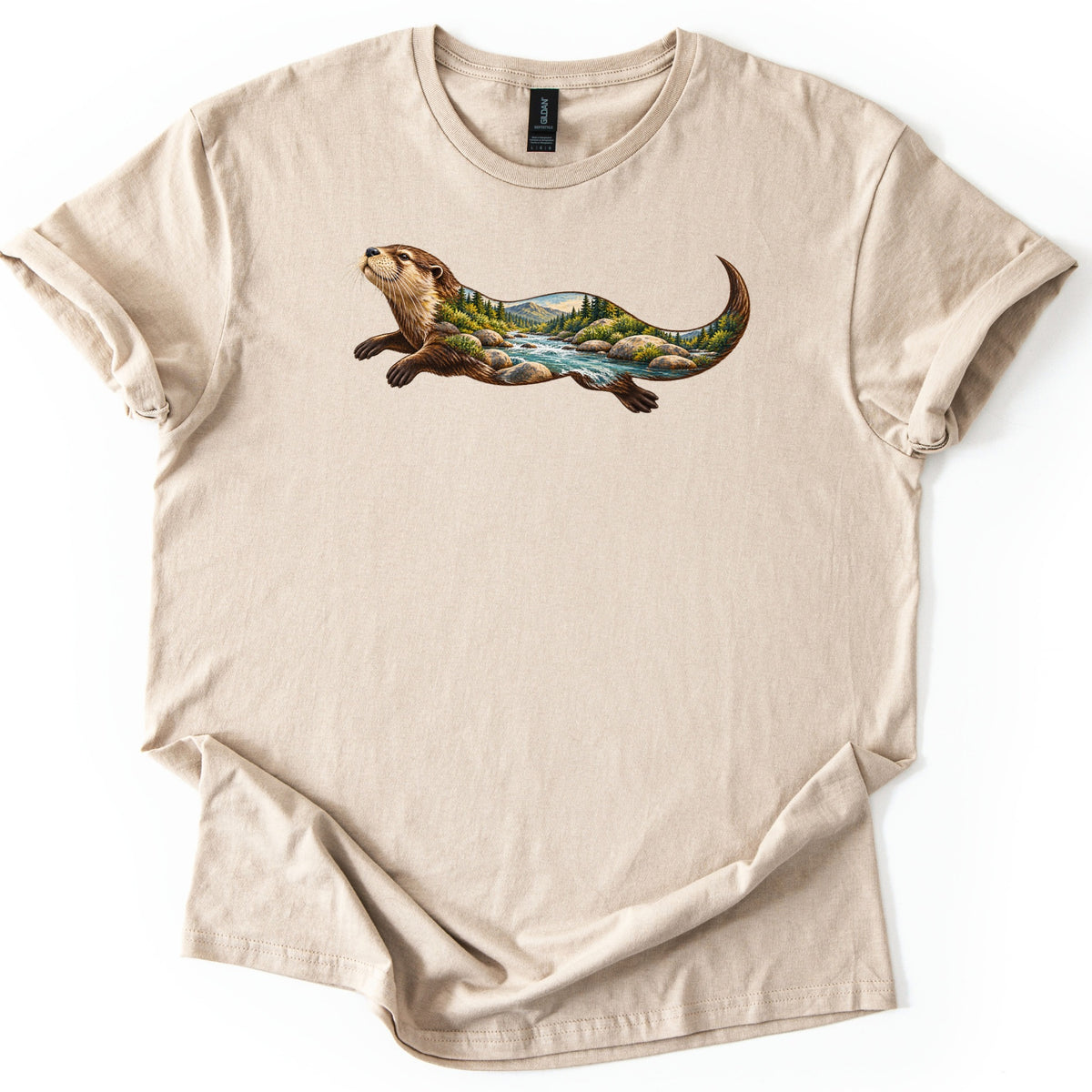 Otter River Bend Comfort Colors T-shirt in Sand, laid flat. A river otter stretches forward with its head raised, its body filled with a stream rushing over boulders, pine trees and distant mountains. No words. Sleeves are cuffed and the hem folded on white.