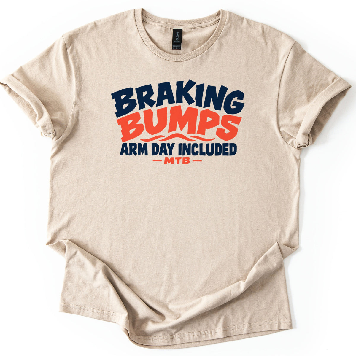Braking Bumps Arm Day Included Comfort Colors T-shirt in Sand, laid flat. Wavy lettering reads Braking in navy and Bumps in orange over orange wave lines, then Arm Day Included in navy and MTB in orange between dashes. The shirt has rolled sleeves on a white background.