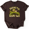 Taking the Scenic Inch Comfort Colors T-shirt in Dark Chocolate, laid flat. A spiral-shelled snail crawls along a moss-covered log with small rocks, between the moss-dripping words Taking the Scenic Inch. The shirt has rolled sleeves on a plain white background.