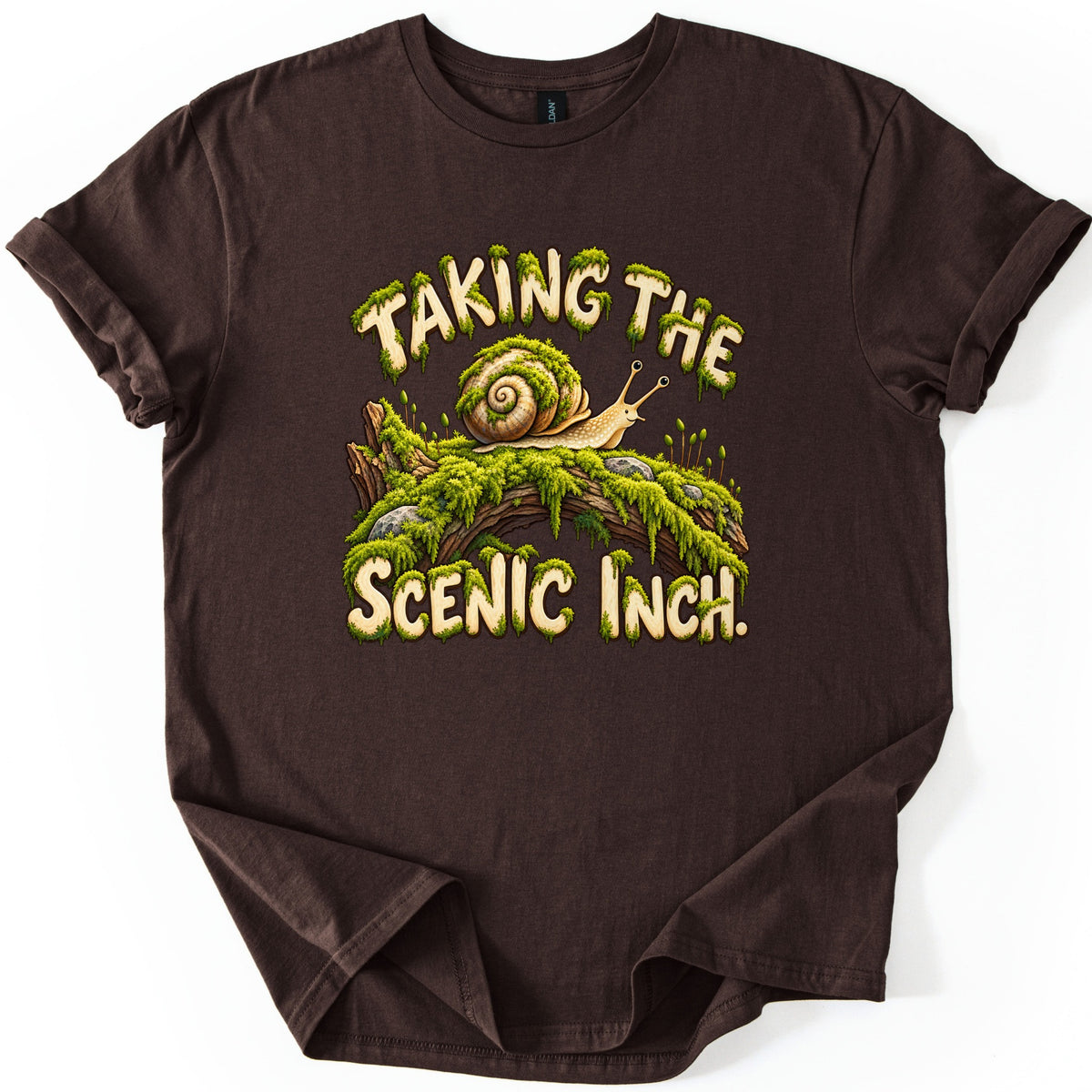 Taking the Scenic Inch Comfort Colors T-shirt in Dark Chocolate, laid flat. A spiral-shelled snail crawls along a moss-covered log with small rocks, between the moss-dripping words Taking the Scenic Inch. The shirt has rolled sleeves on a plain white background.