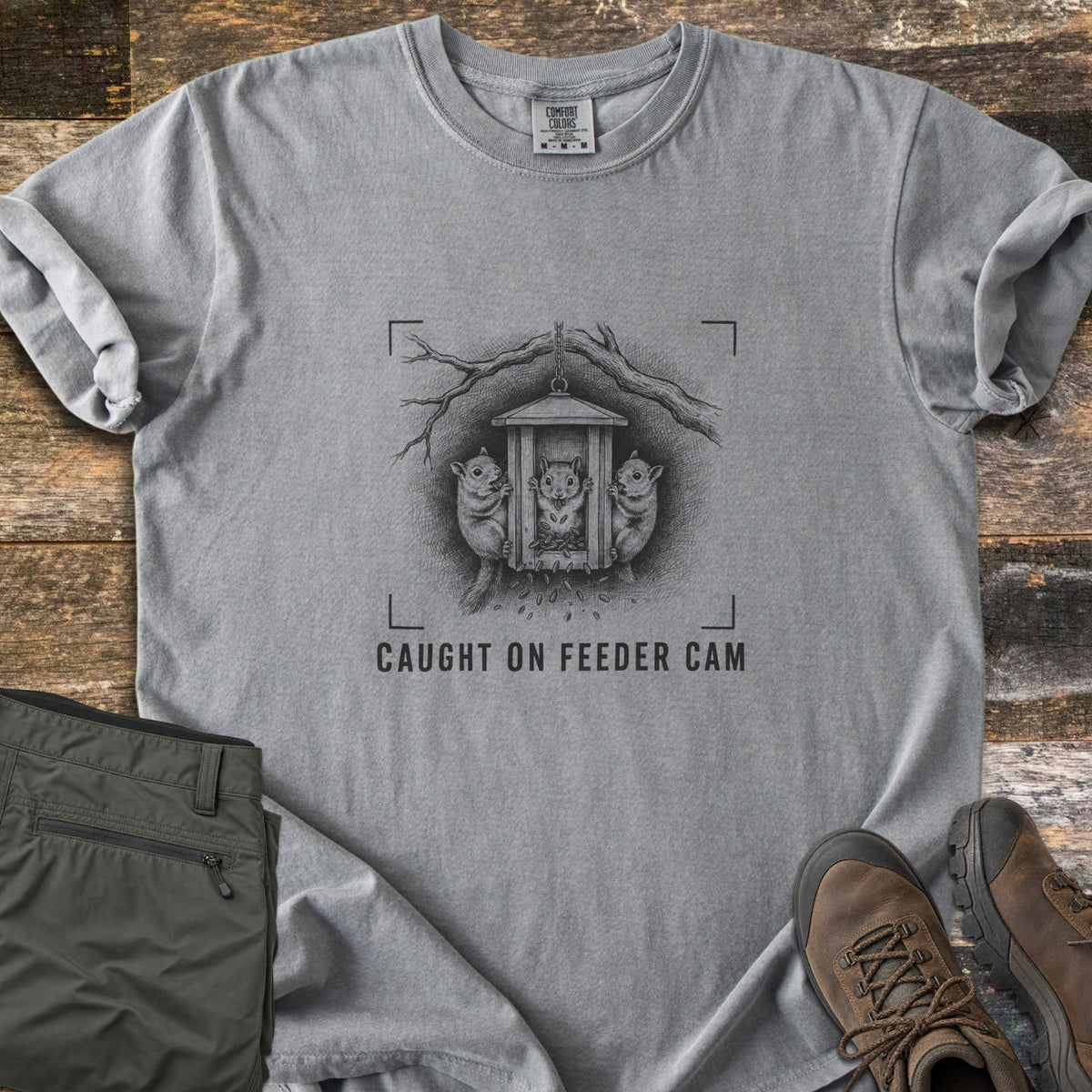 Caught On Feeder Cam Comfort Colors T-shirt in Grey, laid flat. A black ink sketch of three squirrels raiding a hanging wooden feeder inside camera viewfinder corners, above the words CAUGHT ON FEEDER CAM. Folded olive pants and brown hiking boots sit on weathered wood planks.