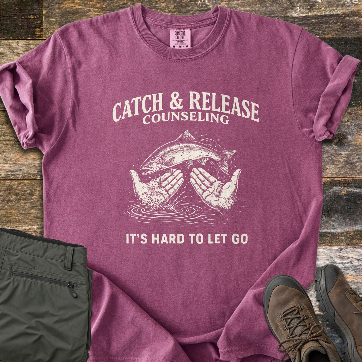 Catch & Release Counseling Comfort Colors T-shirt in Berry, laid flat. A trout leaps between two open hands over rippling water under the words Catch & Release Counseling, with It's Hard to Let Go below, in cream ink. Folded olive hiking pants and brown boots sit on weathered wood.