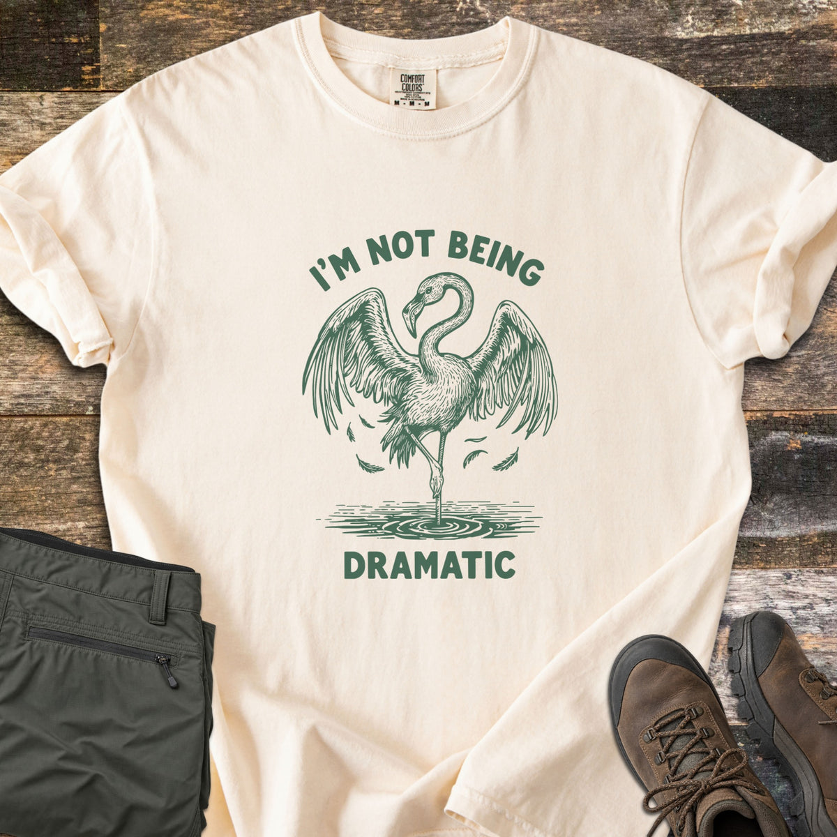 I'm Not Being Dramatic Comfort Colors T-shirt in Ivory, laid flat. A green etched flamingo stands on one leg in rippling water, wings spread and feathers falling, between the words I'm Not Being Dramatic. Olive cargo pants and brown hiking boots sit on a rustic wood floor.