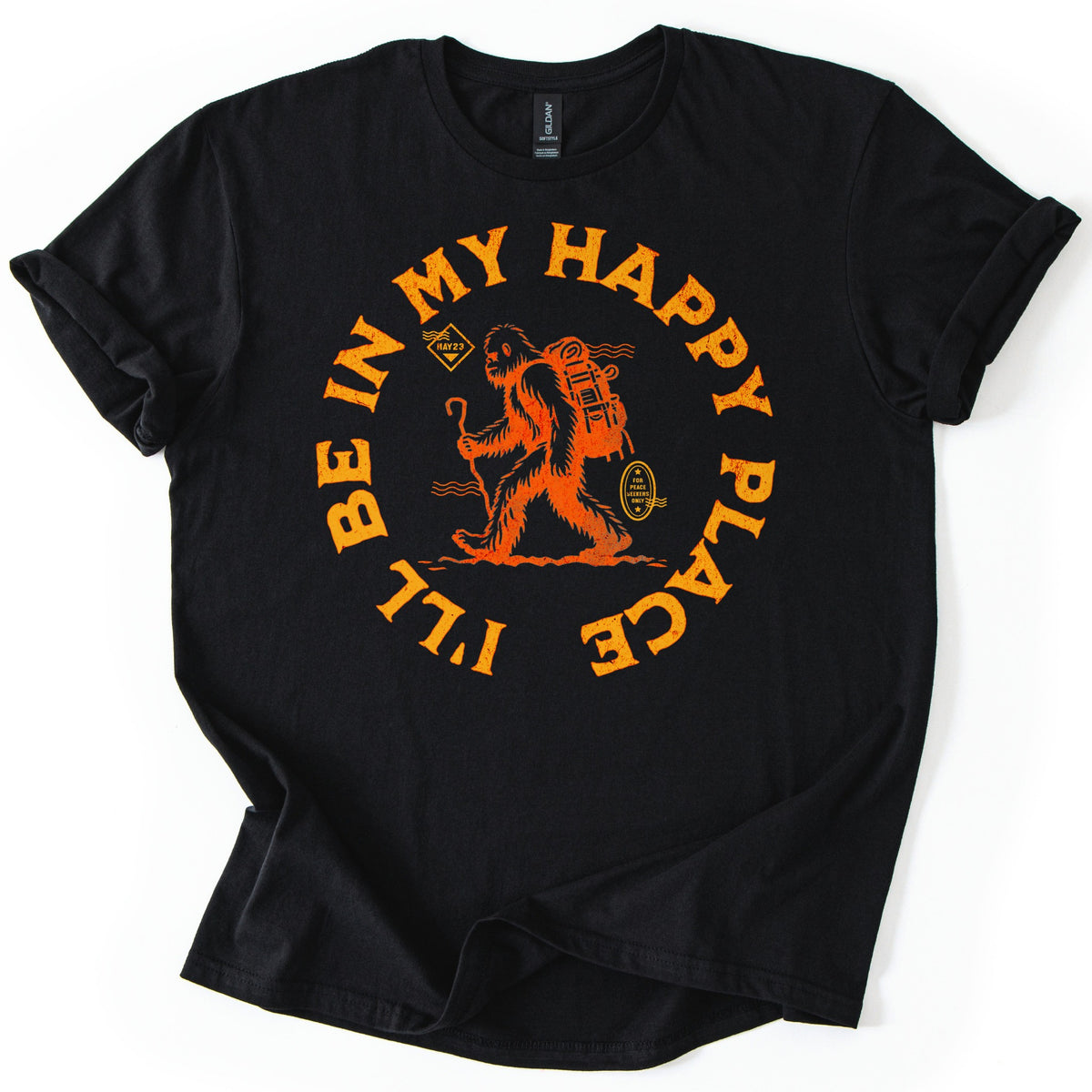 Happy Place Hiking Squatch Comfort Colors T-shirt in Black, laid flat. An orange distressed Bigfoot hikes with a backpack and walking stick, ringed by the curved words I'll Be In My Happy Place, with a small badge reading For Peace Seekers Only. Sleeves are cuffed on a white surface.