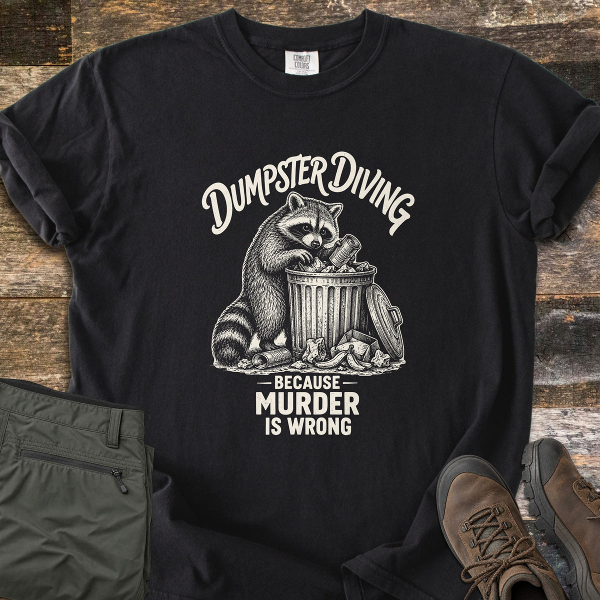 Dumpster Diving Because Murder Is Wrong Comfort Colors T-shirt in Black, laid flat. A sketched raccoon digs through an overflowing metal trash can, with the words Dumpster Diving above and Because Murder Is Wrong below, printed in cream. Olive cargo pants and brown hiking boots lie on a wood floor.