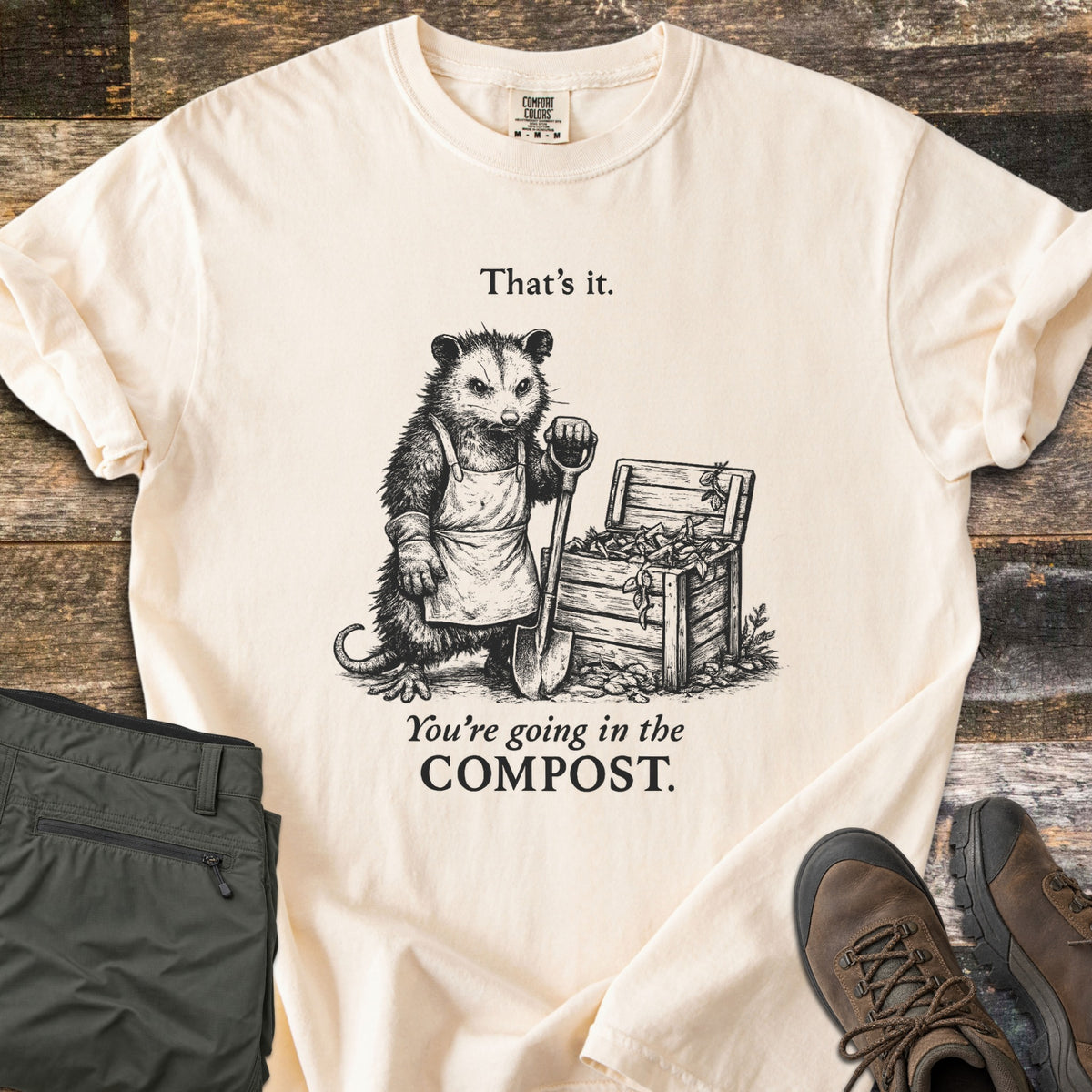 Going In The Compost Possum Comfort Colors T-shirt in Ivory, laid flat. Black ink etching of a scowling possum in an apron, leaning on a shovel by a wooden compost bin, between the words That's it. and You're going in the COMPOST. Olive pants and brown boots lie on wood planks.