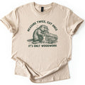 Measure Twice Beaver Comfort Colors T-shirt in Sand, laid flat. A dark green sketch of a beaver gnawing a log beside cut logs, under the arched words Measure Twice, Cut Once and above It's Only Woodwork. The shirt has rolled sleeves on a white background.