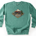 Miles Before Emails Comfort Colors crewneck sweatshirt in Light Green, laid flat. A diamond-shaped embroidered-style patch shows a red ATV on rocks with pine trees and mountains behind it, above the words MILES BEFORE EMAILS. The sweatshirt lies on a white surface.