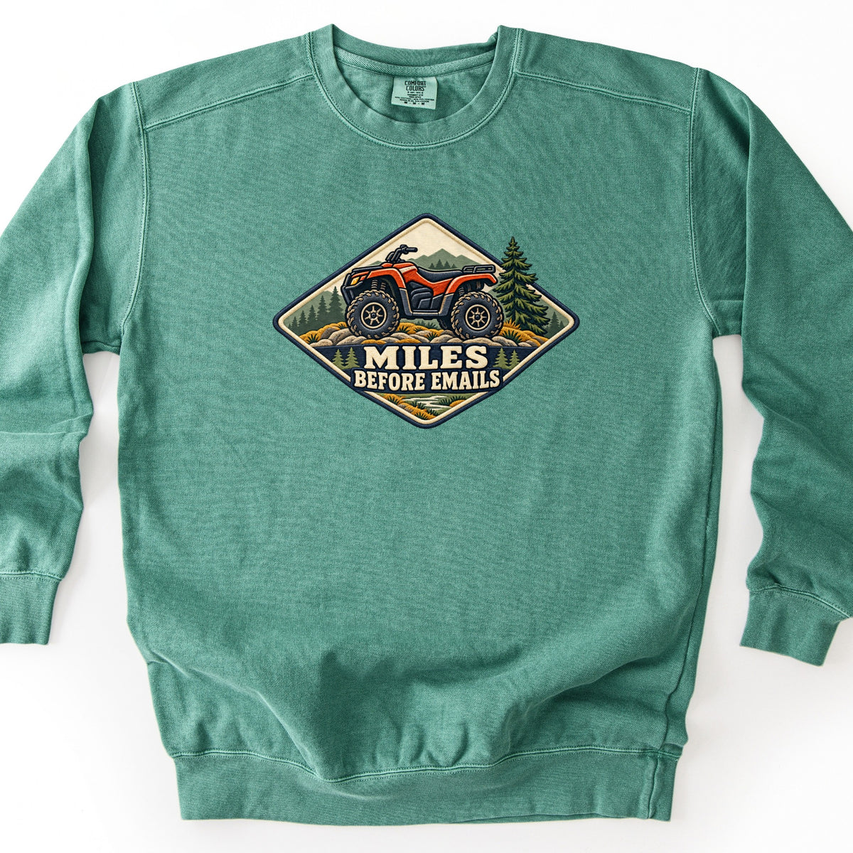 Miles Before Emails Comfort Colors crewneck sweatshirt in Light Green, laid flat. A diamond-shaped embroidered-style patch shows a red ATV on rocks with pine trees and mountains behind it, above the words MILES BEFORE EMAILS. The sweatshirt lies on a white surface.