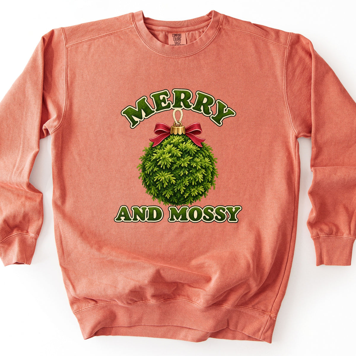 Merry and Mossy Comfort Colors crewneck sweatshirt in Terracotta, laid flat. A round green moss ball ornament with a gold cap, loop, and red bow, between the words Merry and And Mossy in green arched lettering. Shown on a white background.
