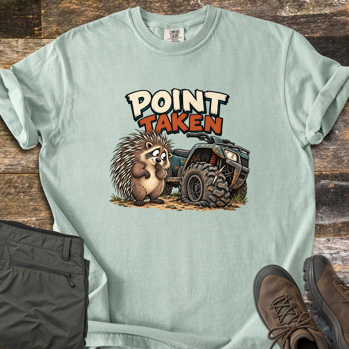 Point Taken Comfort Colors T-shirt in Bay, laid flat. A cartoon porcupine looks sheepishly at a muddy ATV with a flat front tire pierced by a quill, under the words Point Taken. Folded olive pants and brown hiking boots sit beside it on weathered wood.