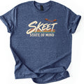 Skeet State of Mind Comfort Colors T-shirt in Heather Navy, laid flat. The word SKEET in cream brushstroke letters over orange, gold and teal stripes, with two orange clay targets flying above and STATE OF MIND below. Sleeves are cuffed on a white background.