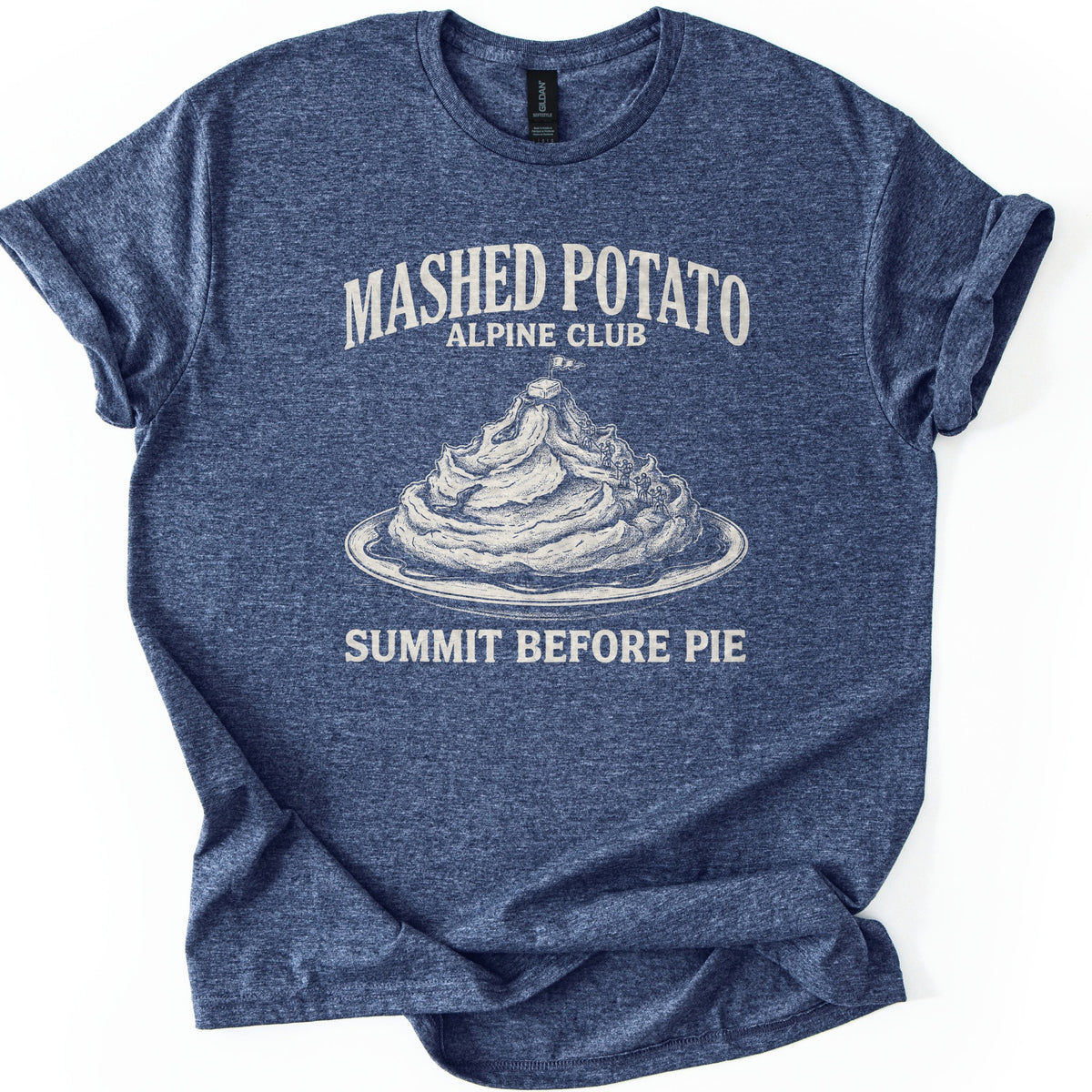 Mashed Potato Alpine Club Comfort Colors T-shirt in Heather Navy, laid flat. Cream etched art of a mashed potato mountain on a plate, with tiny climbers and a butter pat and flag on top, under Mashed Potato Alpine Club and above Summit Before Pie. Rolled sleeves, white background.