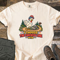 I Choose the Chicken Line MTB Comfort Colors T-shirt in Ivory, laid flat. A cartoon chicken in a bike helmet stands on a dirt trail by rocks, pine trees and a signpost above the words I Choose the Chicken Line MTB. Olive pants and brown hiking boots lie beside it on wood.