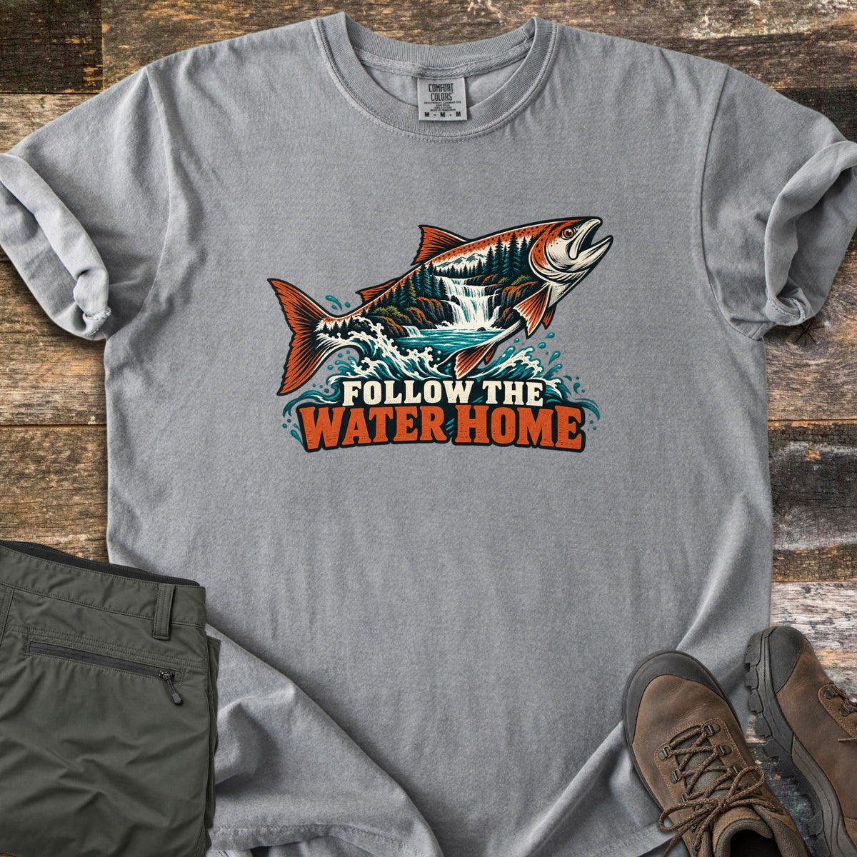 Follow the Water Home Comfort Colors T-shirt in Grey, laid flat. A salmon leaps from splashing water, its body filled with a forest and waterfall scene, above the words Follow the Water Home. Laid on wood planks beside folded olive hiking pants and brown hiking boots.