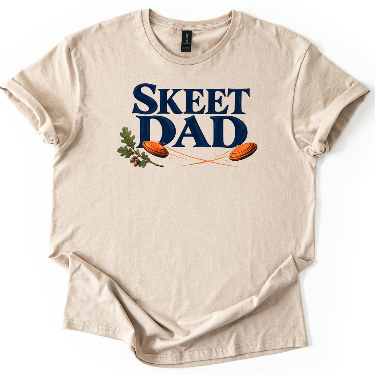 Skeet Dad Comfort Colors T-shirt in Sand, laid flat. The words SKEET DAD in large navy letters, with an oak sprig and acorns on the left and two orange clay targets flying with crossing orange trails. Sleeves are cuffed, on a white background.