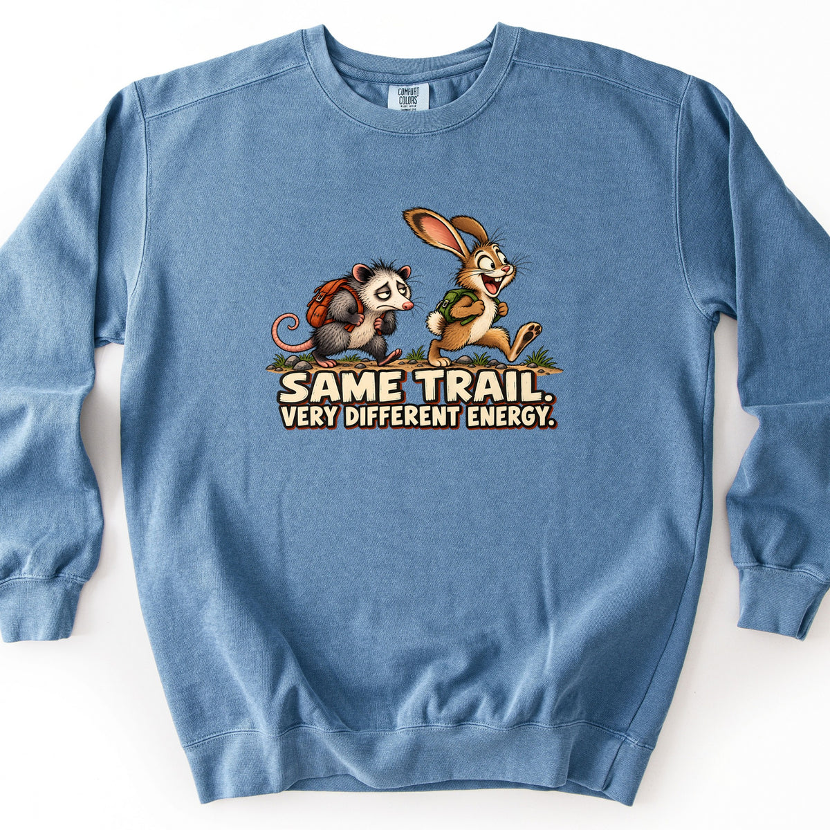 Same Trail. Very Different Energy. Comfort Colors crewneck sweatshirt in Blue Jean, laid flat. A weary cartoon opossum with an orange backpack trudges behind a grinning rabbit with a green backpack on a dirt trail, above the words Same Trail. Very Different Energy. Shown on a white surface.